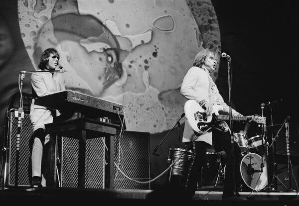 A black-and-white image of two men playing instruments on a stage, with a psychedelic light-show backdrop behind them.