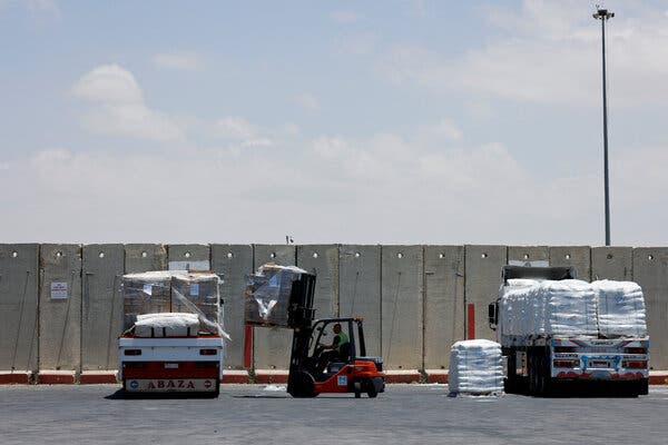 Two trucks and a forklift carrying sacks and pallets of goods, with a concrete wall beyond them.