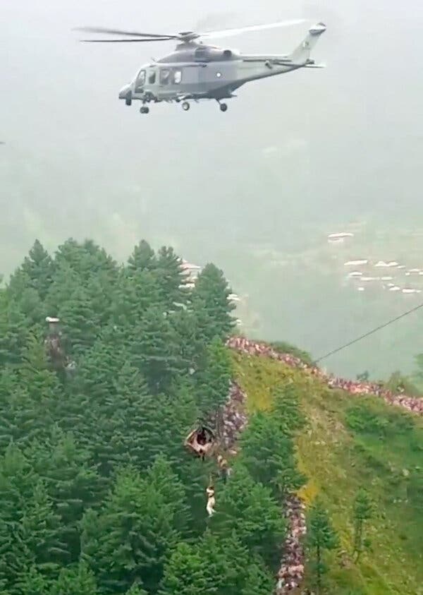 A helicopter hovers over a wooded valley with a person dangling from a rope beneath a stranded cable car.