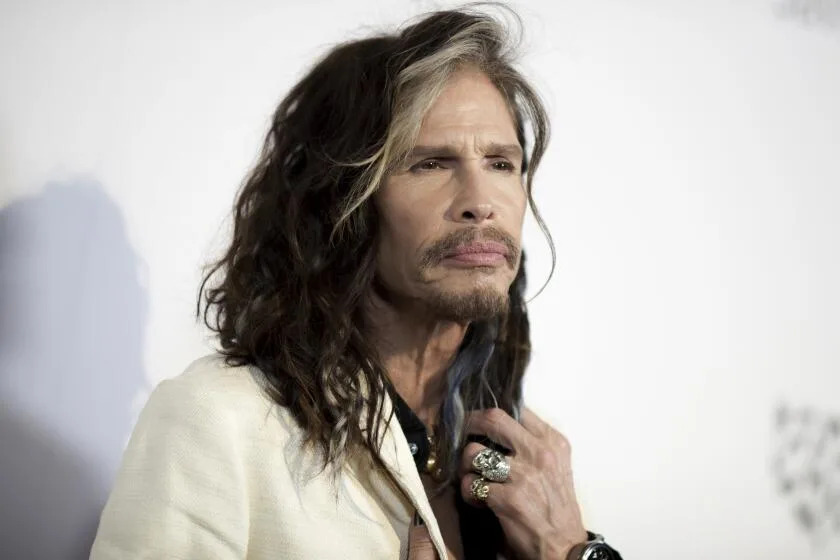 Steven Tyler is posing with his hand with rings is at his neck as his hair flows down toward his cream suit jacket