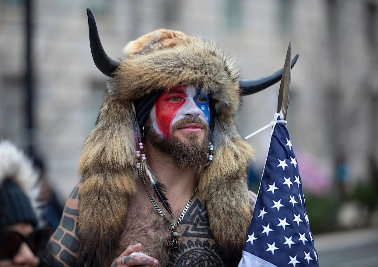 A shirtless man with red, white, and blue face paint, a fur head covering with two horns holding an American flag with a spear tip