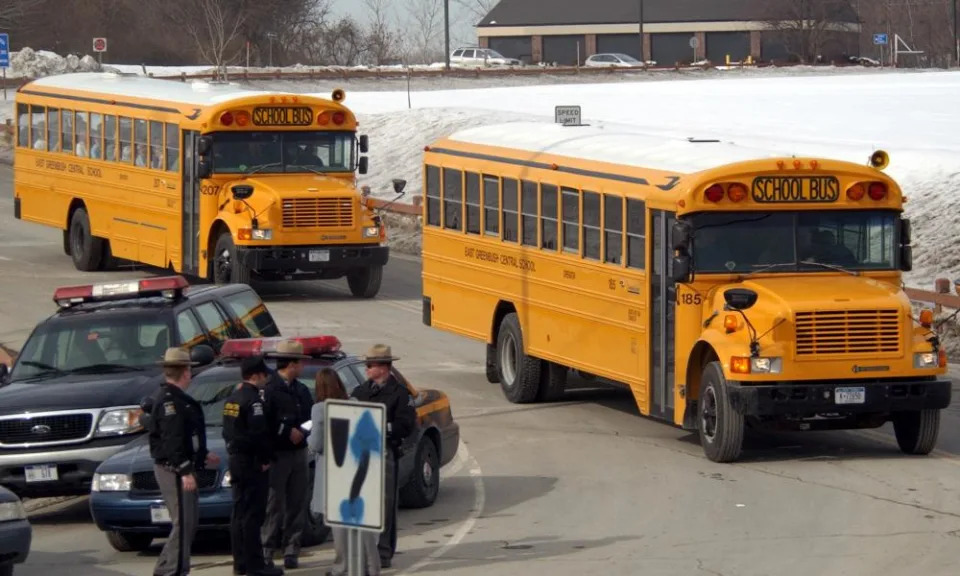 In a parking lot alongside a snowy verge are two orange school buses in a row, next to a sheriff’s SUV and a group of sherrif’s deputies in wide-brimmed hats and winter coats.