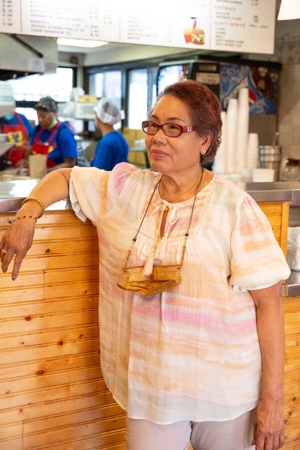 A portrait of an older woman with short brown hair, with her arm resting on the metal counter of a fast food restaurant. She ears glasses and a white shirt horizontally striped with peach and pink brushstrokes of color.