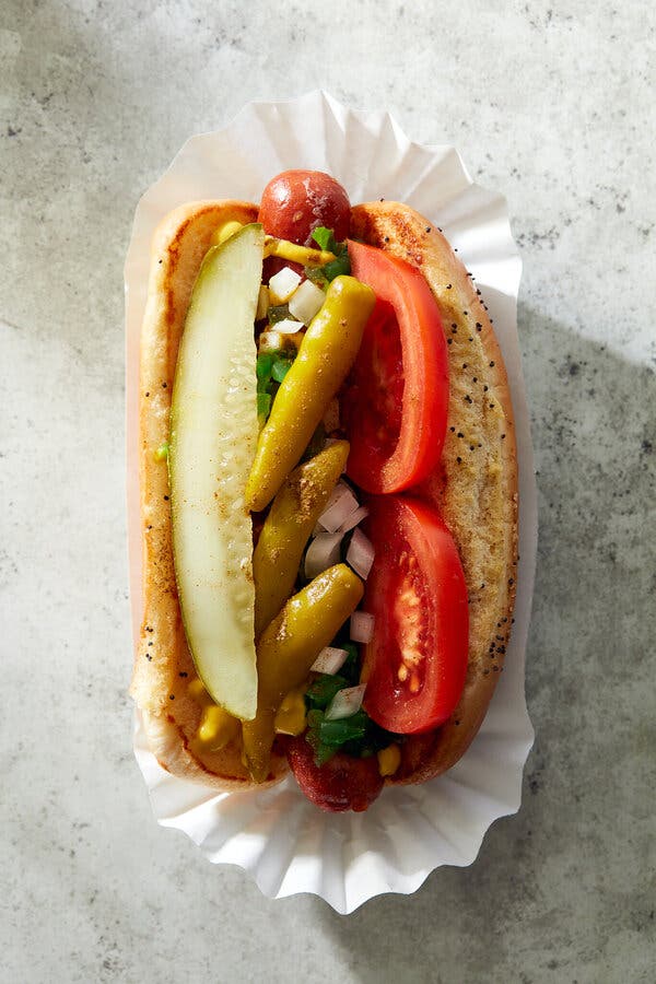 A hot dog in a paper wrapper on a speckled surface. Sitting in a poppy-seed bun, the dog is topped in the Chicago style with a pickle spear, three sport peppers, chopped onions, neon green relish, mustard, two tomato slices and a sprinkle of celery salt.