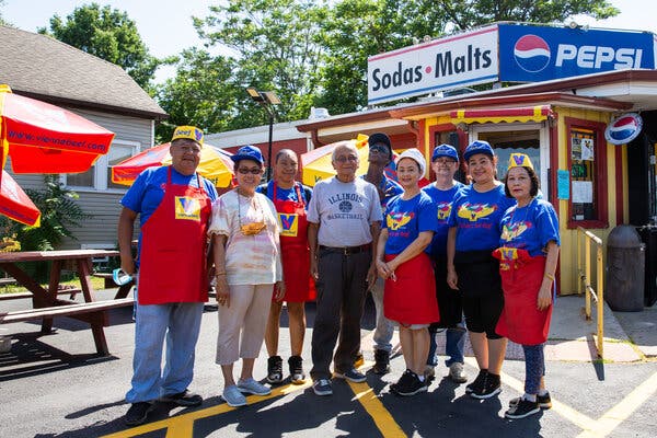 Nine people, many wearing bright blue Vienna Beef hot dog T-shirts, stand in front of a picnic tables with gold and red umbrellas and a restaurant.