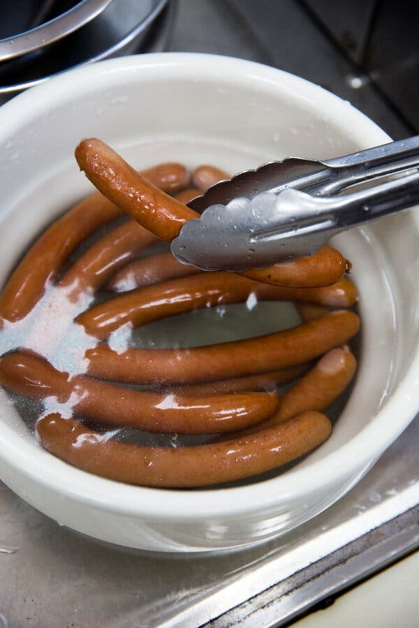 Hot dogs sit in a ceramic crock of oily water. Metal tongs lift one of the brown hot dogs above the container.