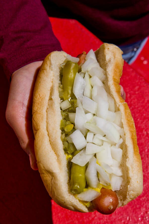 In a closeup photo, a hand holds a hot dog covered in chopped white onions. A bit of yellow mustard, green relish and two sport peppers peek out.