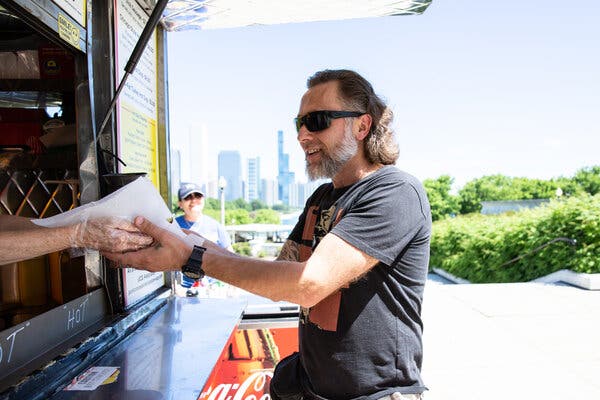 A man in black sunglasses and a black T-shirt with a beard and longish mullet accepts a hot dog from a pair of plastic covered hands within a hot dog cart. The skyline of Chicago is visible in the distance.