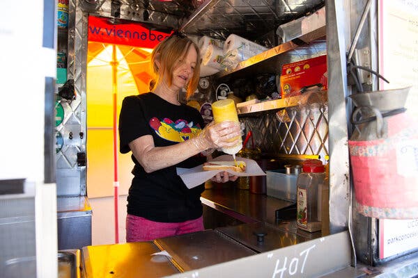 A woman with short dirty blonde hair wearing a black Vienna hot dog T-shirt and pink pants, squirts mustard on a hot dog she is preparing inside a metal street cart.