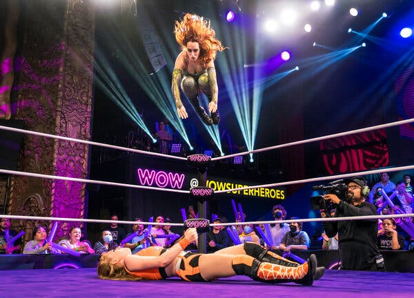 A costumed female wrestler jumps theatrically from the top rope of a wrestling ring toward a female wrestler on the ring floor. The crowd cheers. 