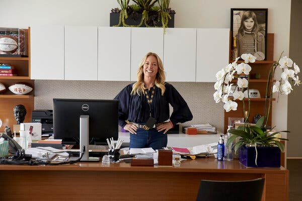 Ms. Buss, wearing jeans and a blue shirt, stands smiling behind an office desk and her hands on her hips.